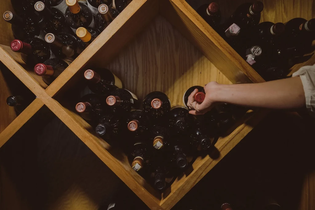 Hand pulling a bottle from a full, dim wooden wine rack.