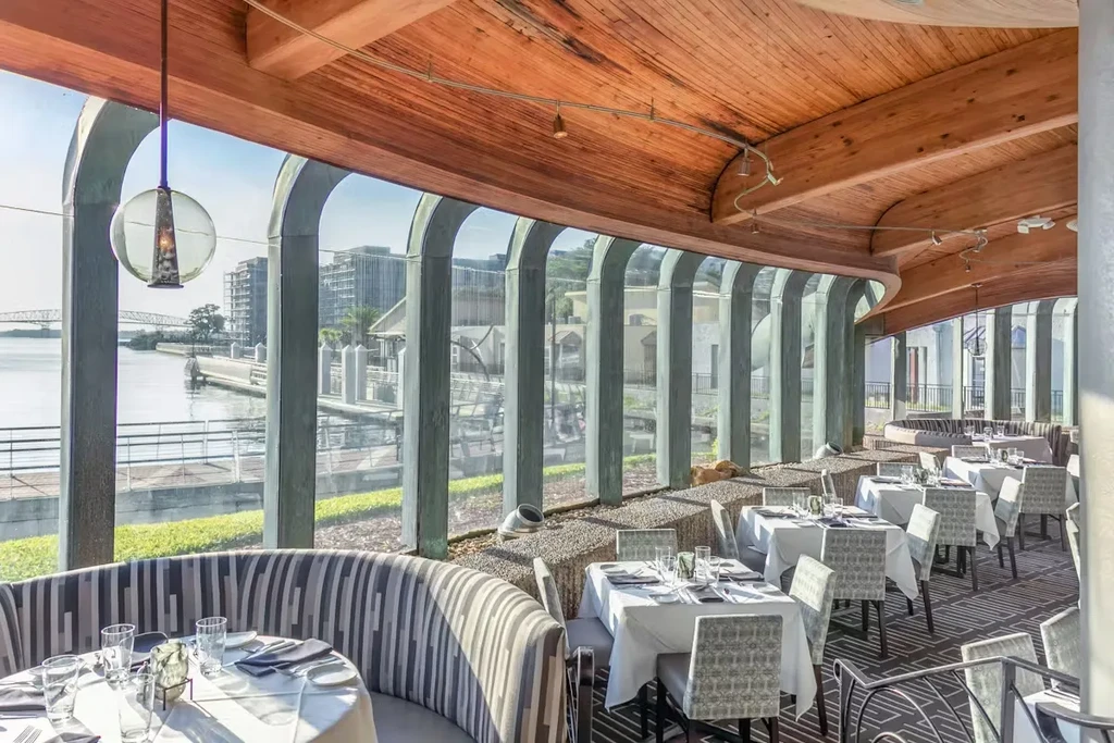 Upscale restaurant interior with curved wooden ceiling, arched windows, water view.