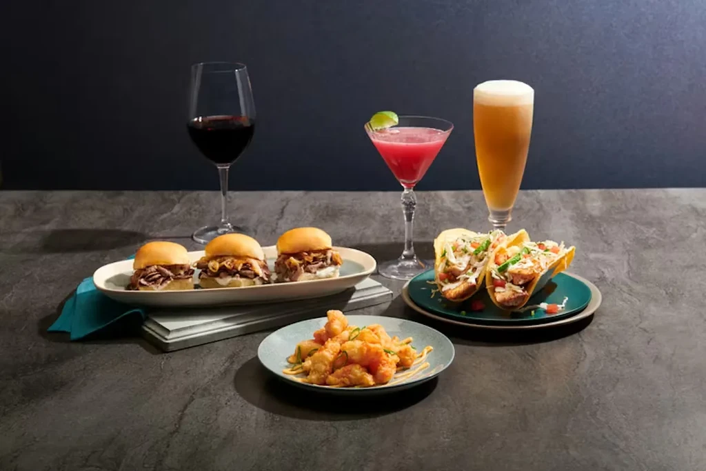 A spread of drinks, sliders, tacos, and fried food on a dark table.