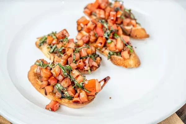 Four bruschetta with tomatoes, basil, and balsamic glaze on a white plate.
