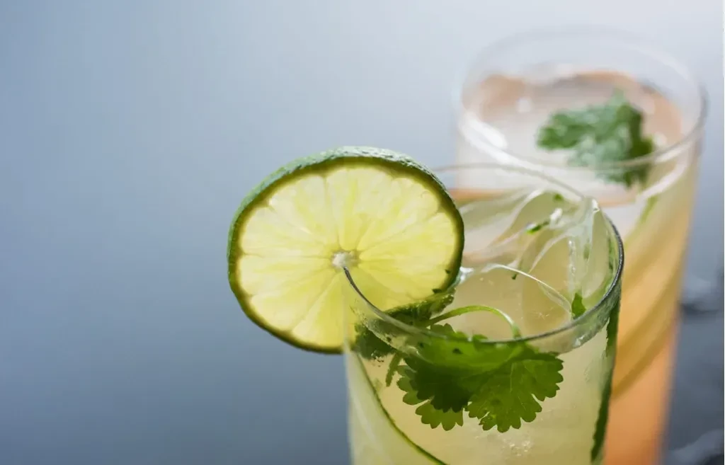 Yellow-green drink in glass with ice, green herbs, thin strips, lime slice.