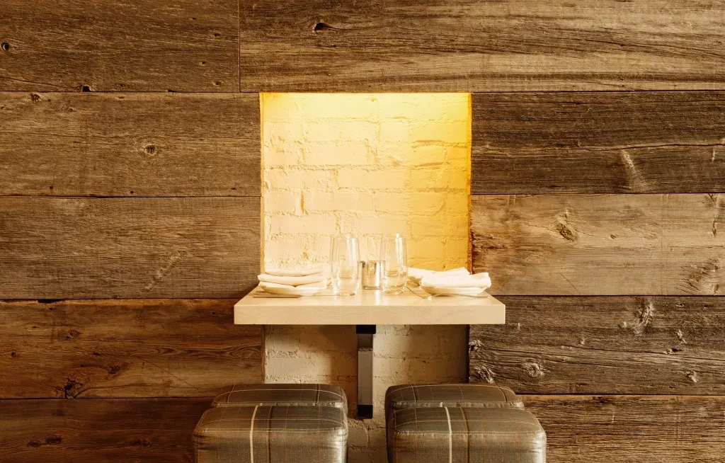 Dining nook: rustic wood wall, brick alcove, shelf, glasses, two stools.