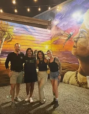 Smiling group of four stands by a tropical beach and face mural.