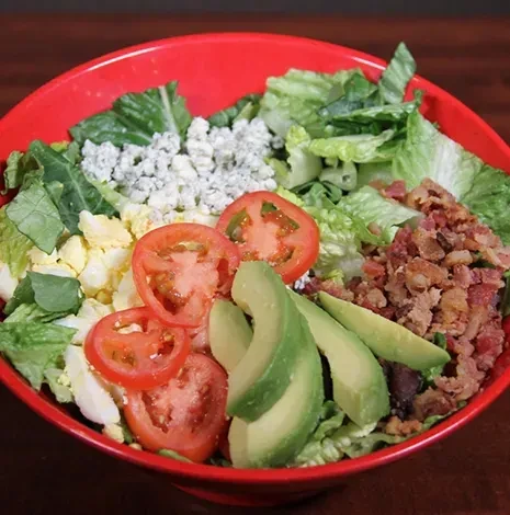 Vibrant salad with avocado, bacon, egg, and blue cheese in red bowl.