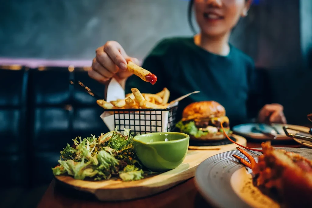 Hand holds ketchup-dipped fry from basket, next to burger, salad.