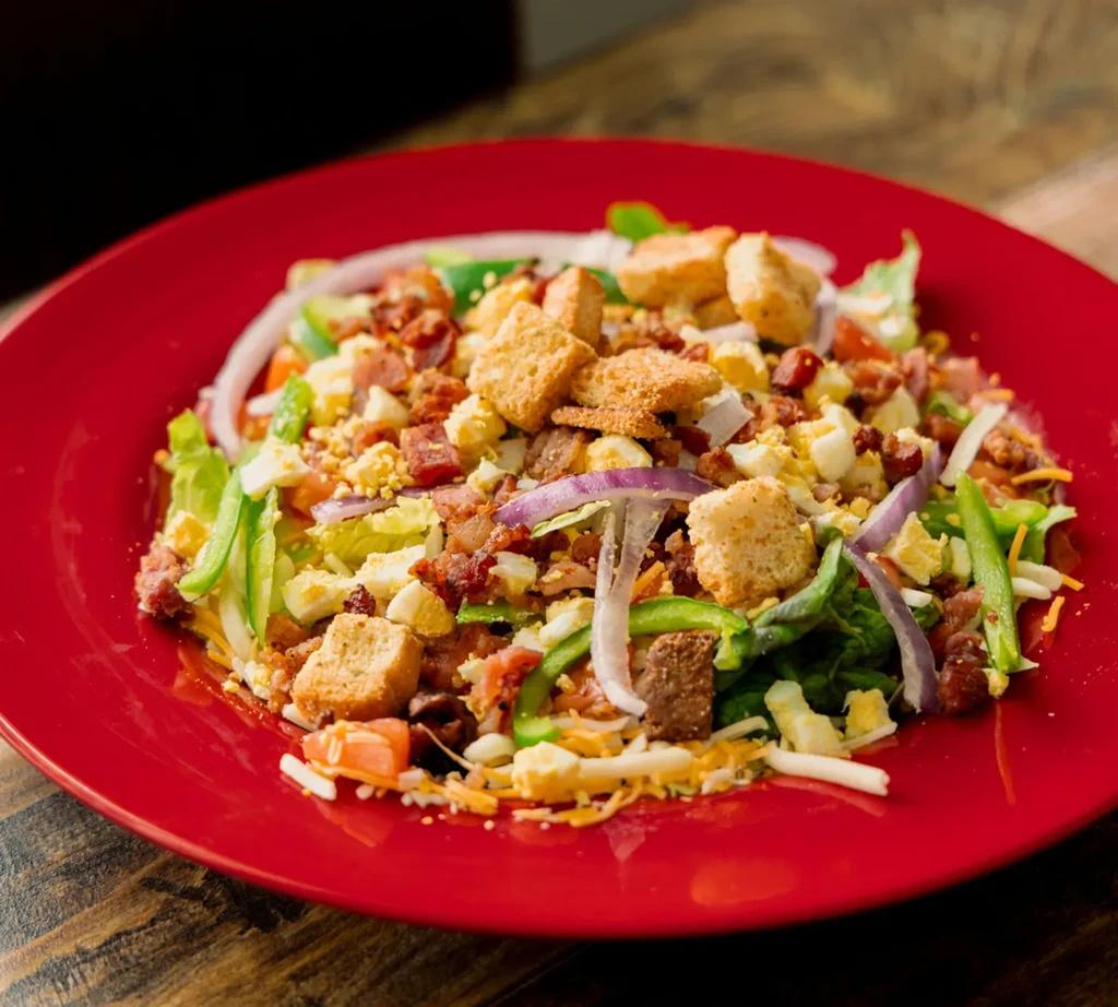 Large colorful salad with lettuce, egg, bacon, croutons on red plate.