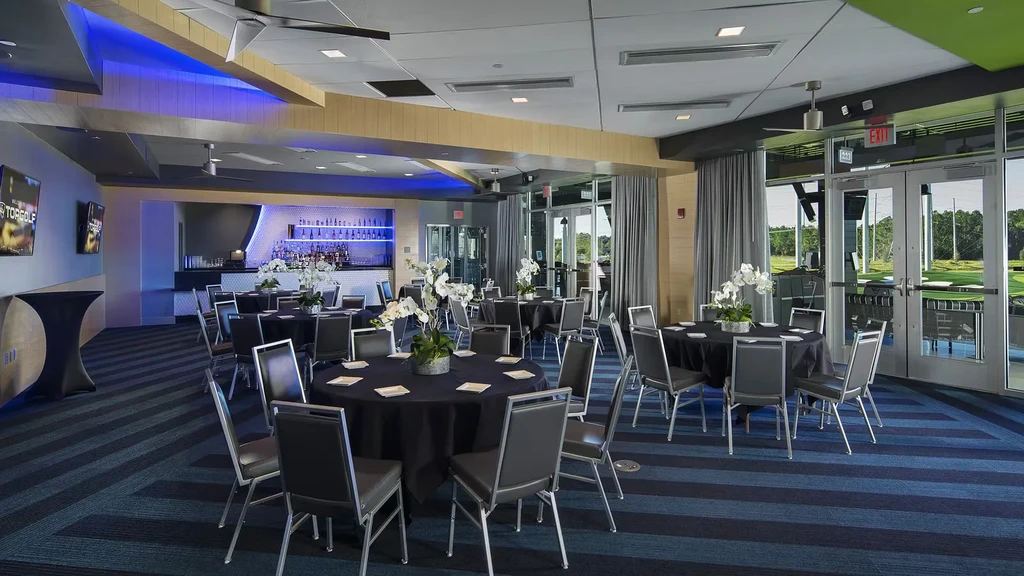 Modern TopGolf venue with tables, blue-lit bar, golf course view.