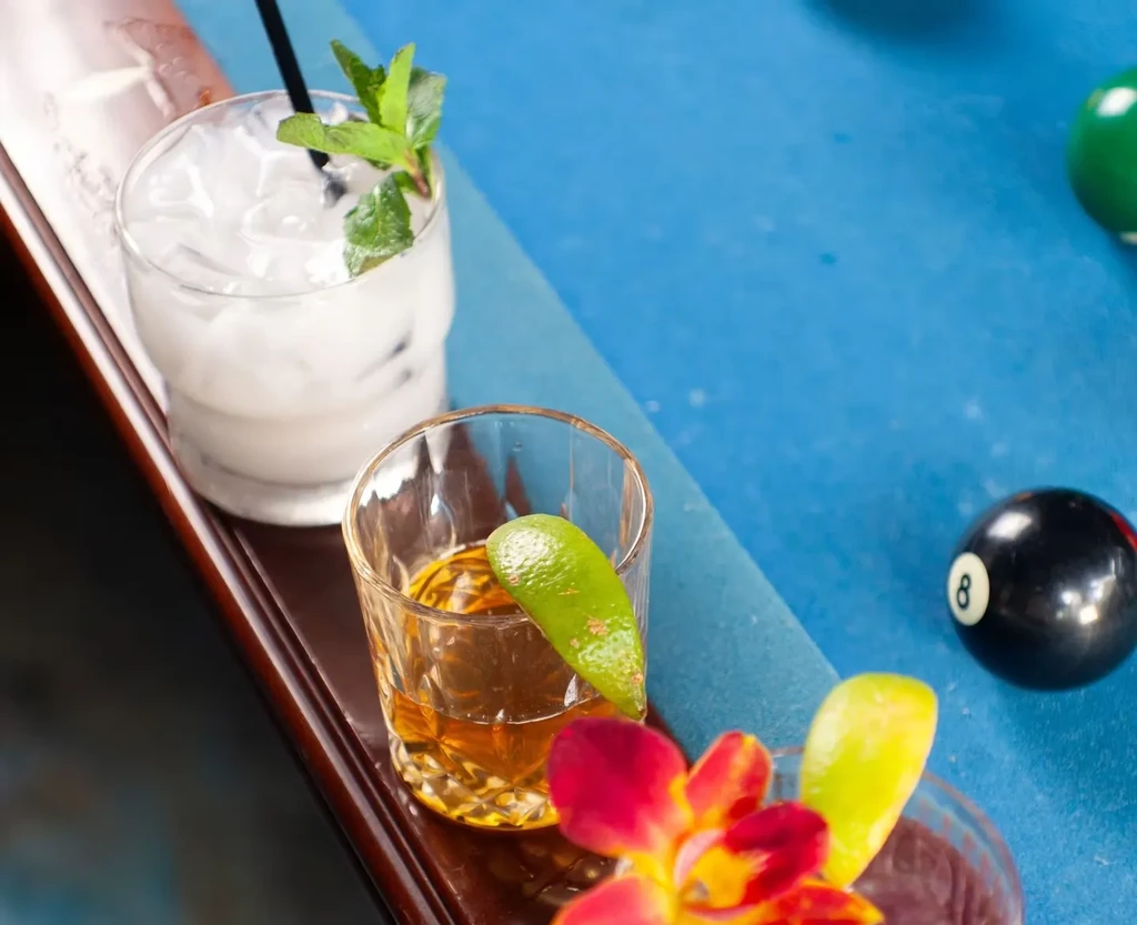 Two garnished drinks, pool balls, and a flower on a wooden surface.