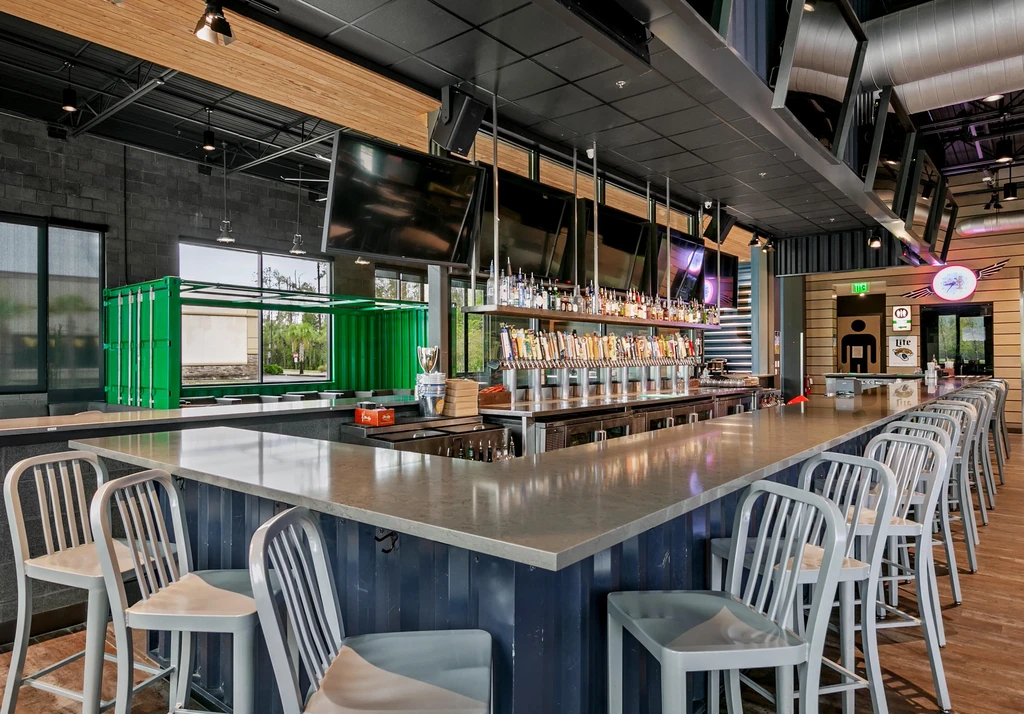 Modern industrial bar interior, L-shaped counter, numerous taps, empty stools.