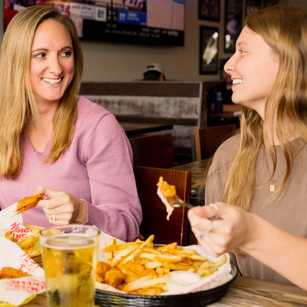 Two women smiling, eating chicken and fries at a sports bar.
