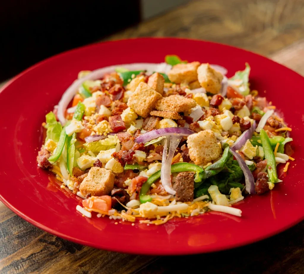 Hearty salad on red plate with bacon, egg, cheese, and croutons.