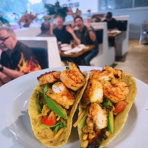 Grilled shrimp tacos on a plate, blurred restaurant with waving patrons.