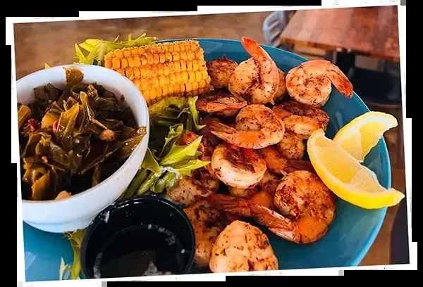 Grilled shrimp, collard greens, corn, and lemon wedges on a blue plate.