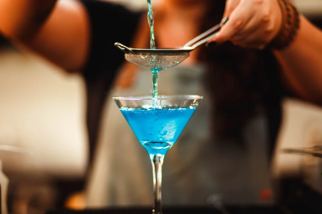 Hands straining blue liquid into a blue cocktail in a martini glass.
