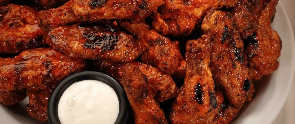 Glossy, grilled chicken wings piled on a white plate with dipping sauce.