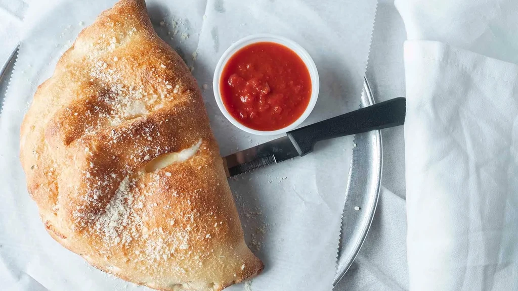 Top-down: Golden-brown savory pastry, marinara sauce, serrated knife.