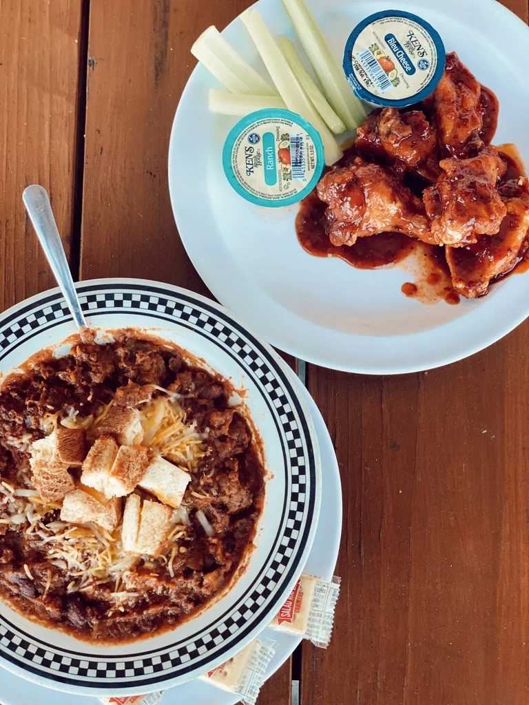Chili and chicken wings with celery and dressings on a rustic table.