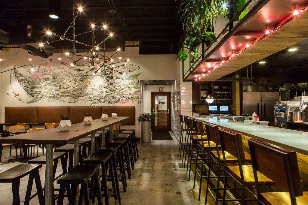 Warmly lit modern restaurant with communal table, bar, and mural.