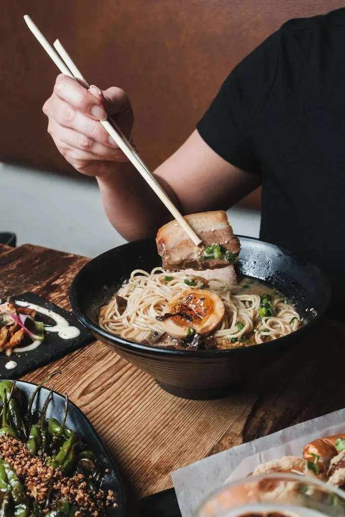 Hands use chopsticks to lift pork belly from a black ramen bowl.