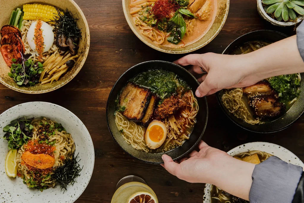 Top-down view of hands placing a ramen bowl among other noodle dishes.
