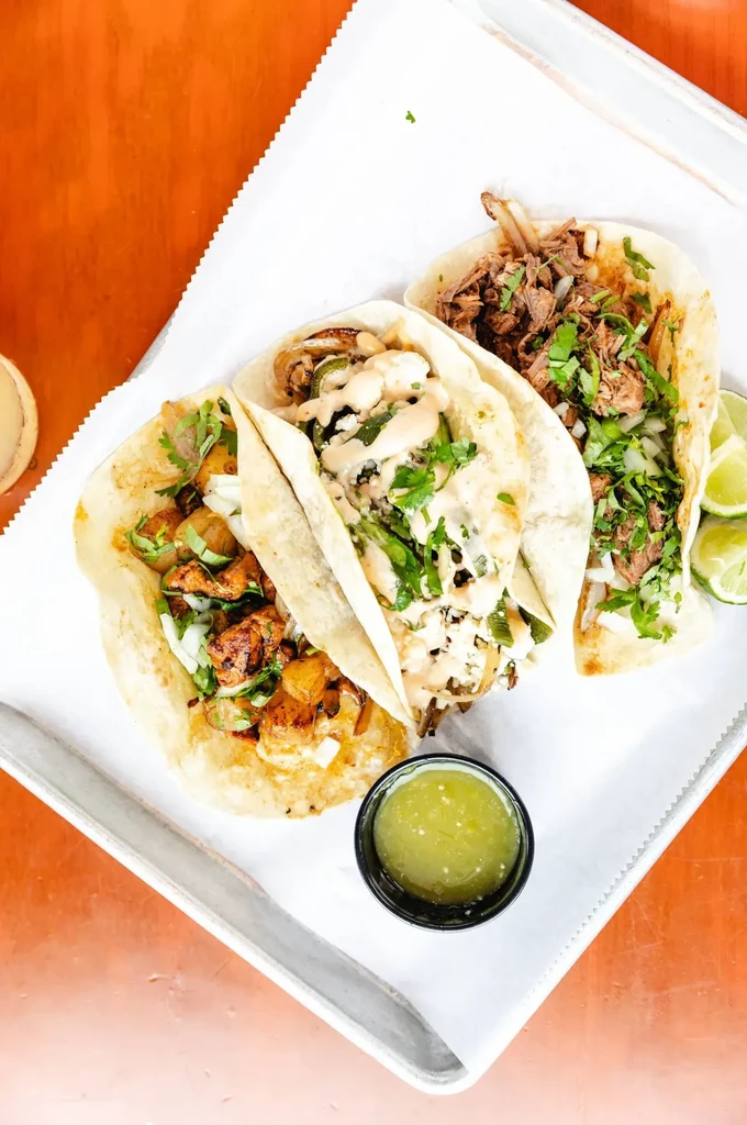 Three tacos with various fillings, salsa, and lime on a tray.