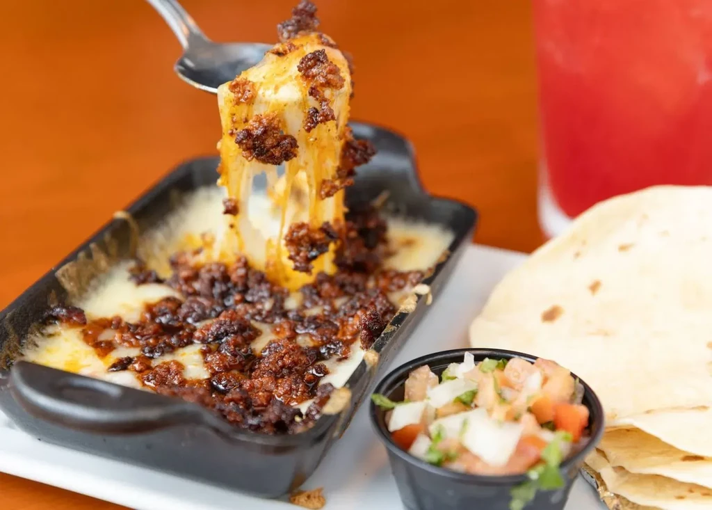 Spoon lifting melted chorizo queso fundido, with pico de gallo and tortillas.