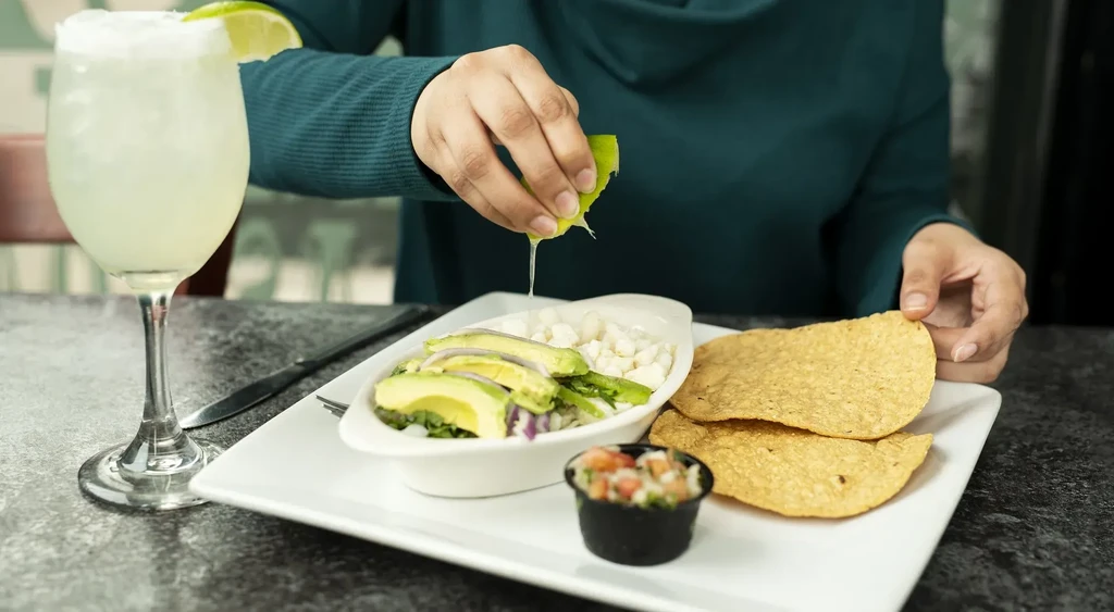 Hands squeeze lime onto ceviche, served with tostadas and a drink.
