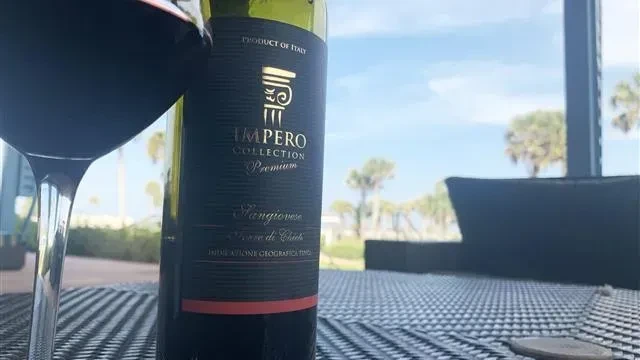 Impero Collection Sangiovese bottle and glass on patio, palm trees.
