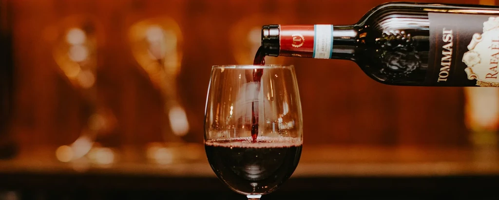Dark red wine pours from a bottle into a glass, blurred warm lights.