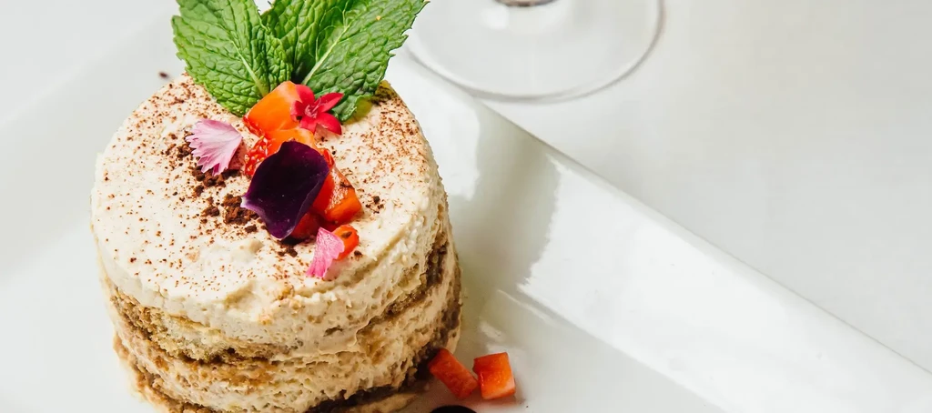 Cylindrical layered dessert with creamy top, cocoa, mint, fruit, and flowers.
