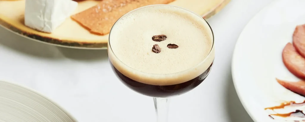 Espresso martini with creamy foam and coffee beans; blurred charcuterie.