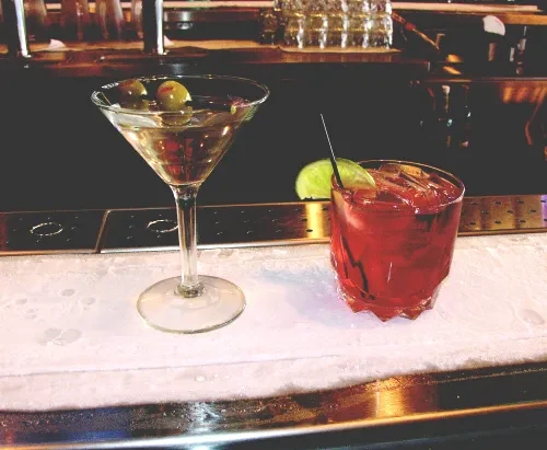 Martini with olives and red cocktail on an iced bar.