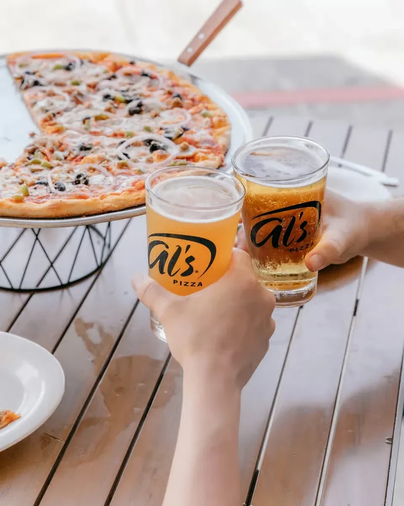 Hands toast Al's PIZZA beers over a pizza with vegetables.