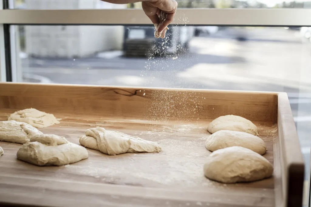 Hand sprinkles flour over unbaked dough portions on a wooden surface.