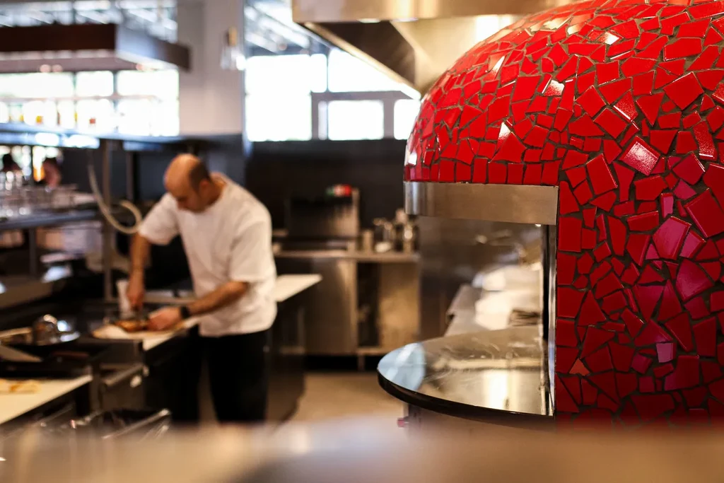 Chef prepares food next to a vibrant red mosaic pizza oven.