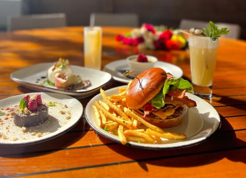 Cheeseburger, fries, chocolate dessert, and burrata on a bright wooden table.