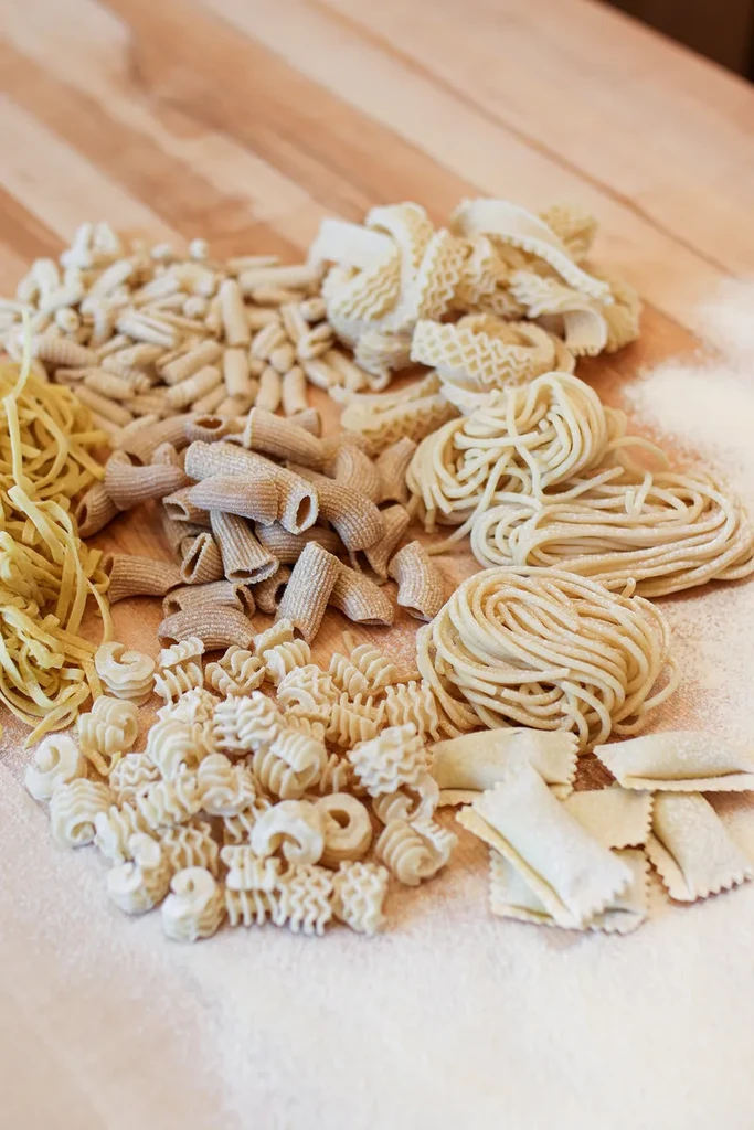 Various raw pasta shapes scattered on a floured wooden surface.