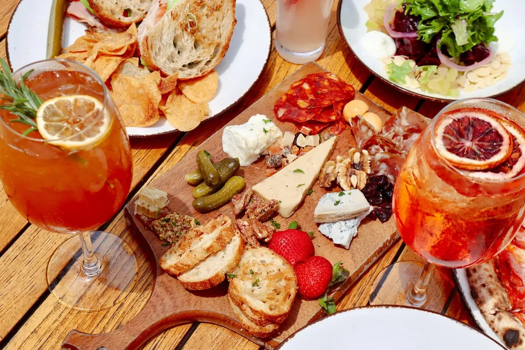 A charcuterie board, sandwiches, salads, and drinks on a wooden table.
