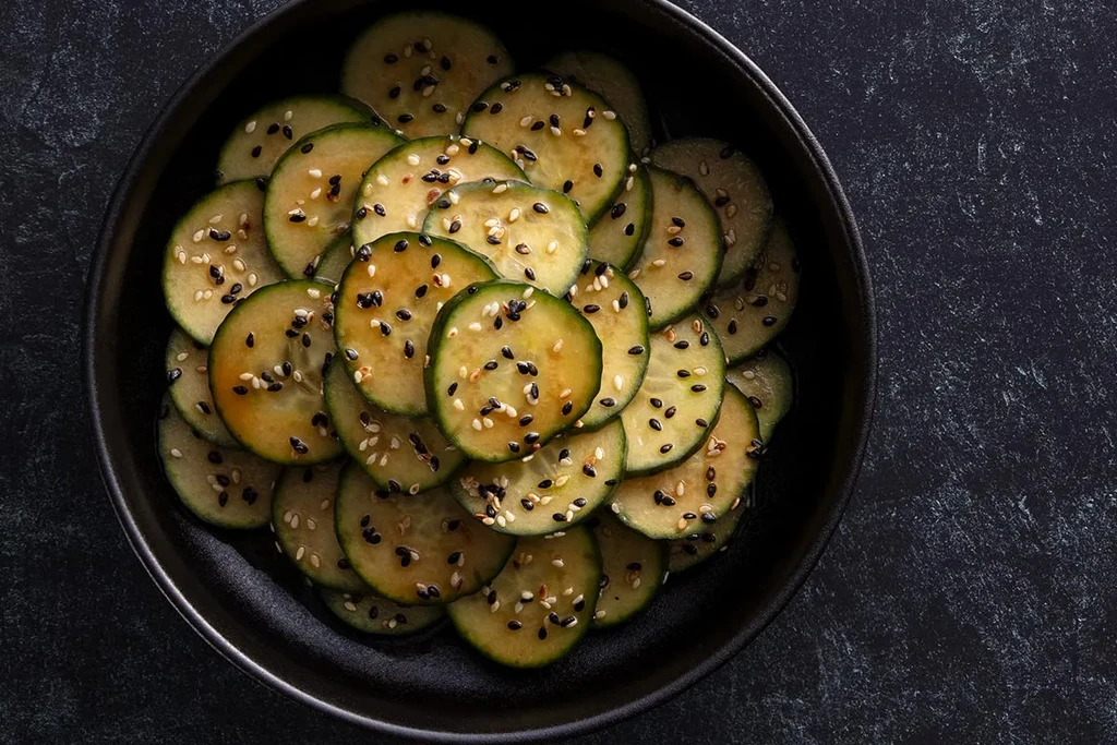 Thinly sliced, marinated cucumbers with sesame seeds in a dark bowl.