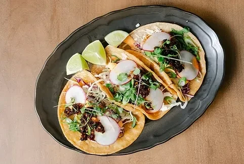 Three tacos with cabbage, radishes, herbs, and lime on a dark platter.
