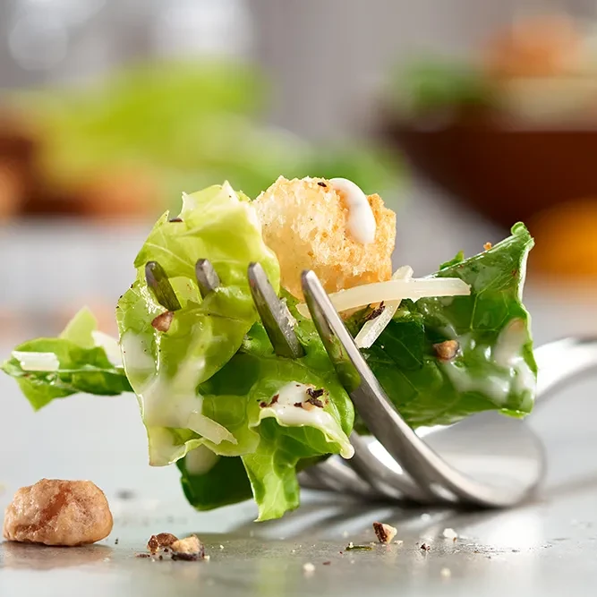 Fork holding a bite of salad with lettuce, crouton, cheese, dressing.