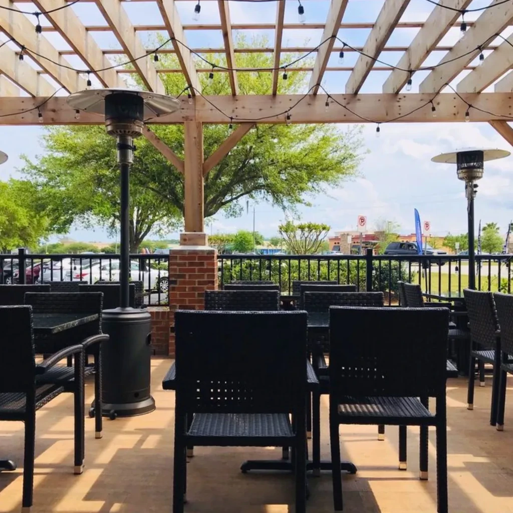 Empty sunlit outdoor patio with dark tables, pergola, and patio heaters.