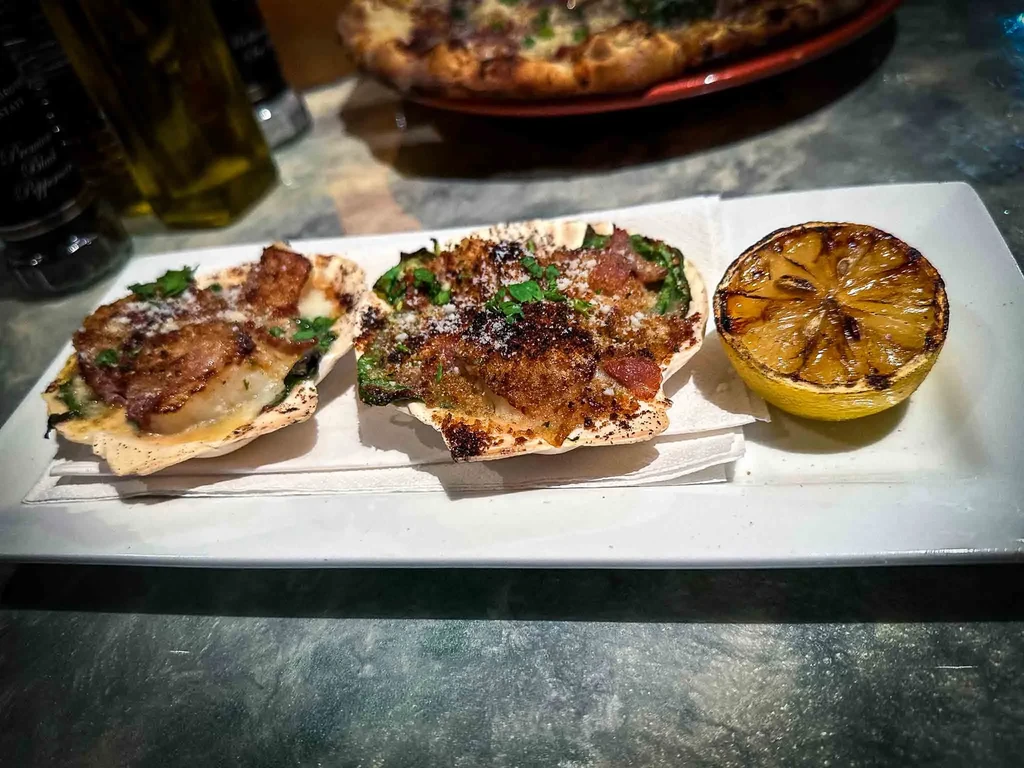 Two gratin-filled scallop shells with herbs and grilled lemon on a plate.