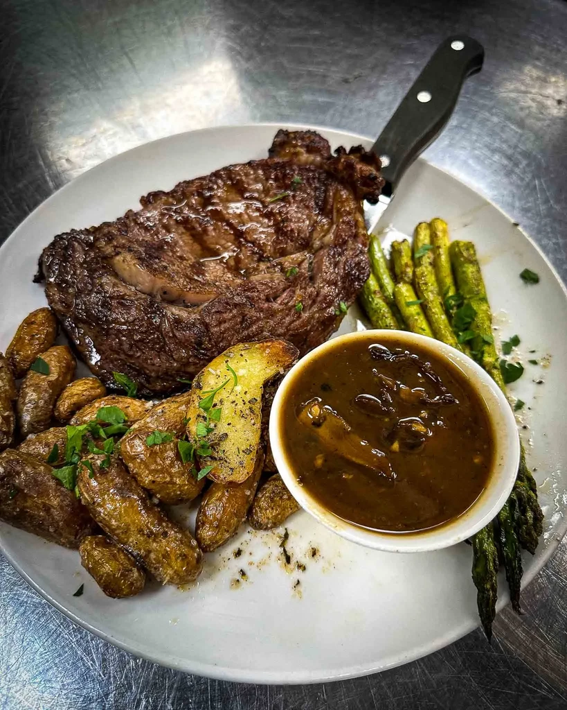 Plated ribeye steak, asparagus, roasted potatoes, dark sauce, and knife.