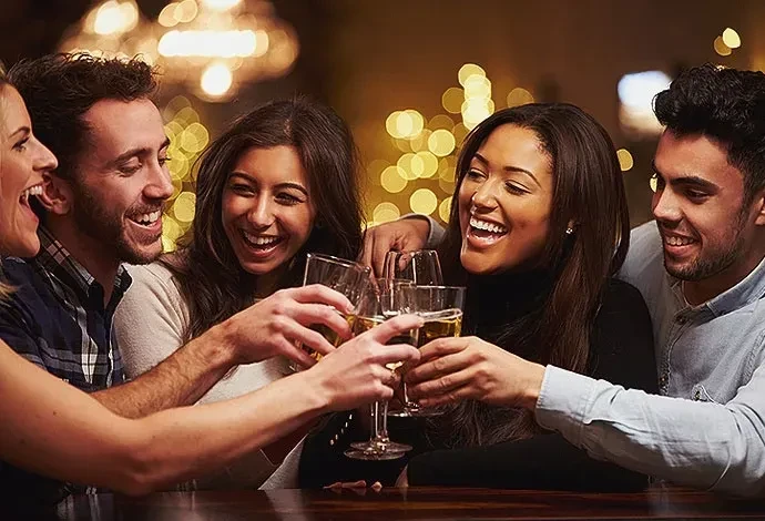 Five smiling adults toasting drinks in a festive blurred background.