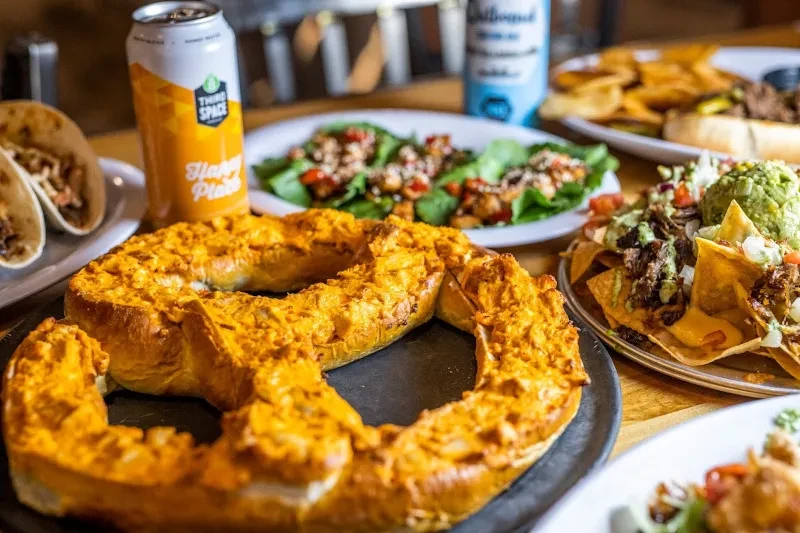Loaded nachos, tacos, baked pretzel, salad, and craft beer on a table.