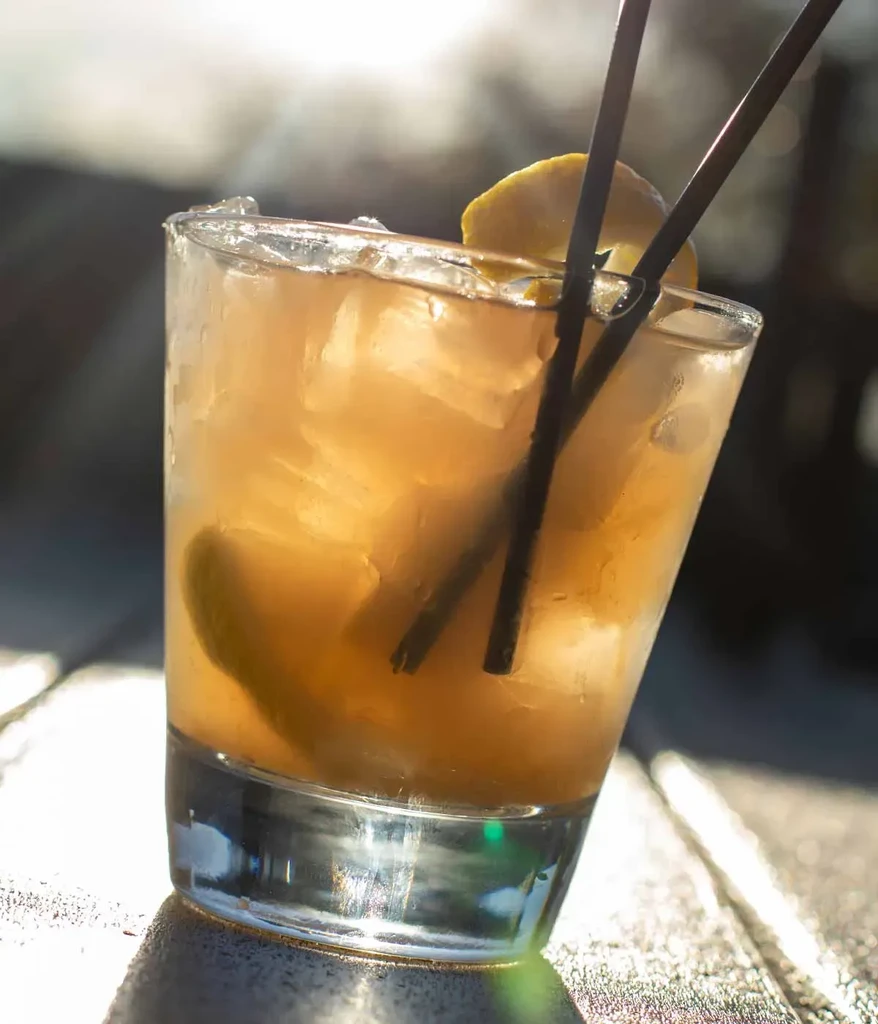 Amber drink with ice, lemon peel, and straws in a glass outdoors.