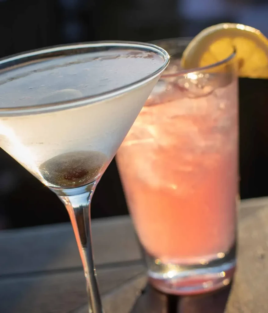 Martini with olive, pink cocktail with lemon slice, on a dark surface.