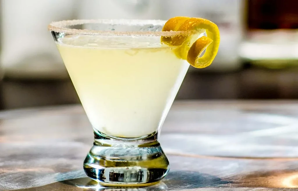 Pale yellow cocktail in martini glass with frosted rim and lemon twist.