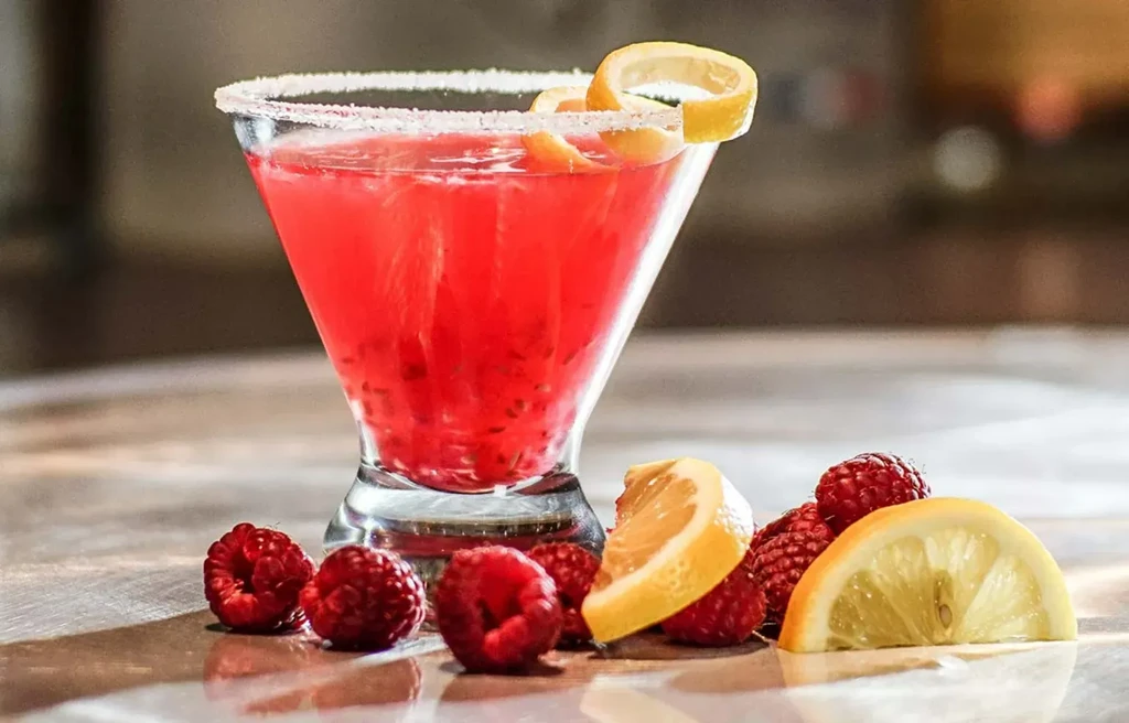 Red cocktail in martini glass with sugared rim, lemon, and raspberries.